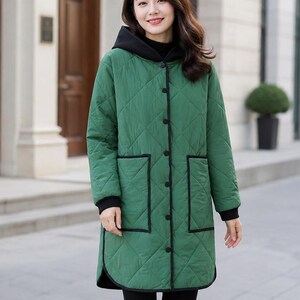 May include: A long-sleeved, knee-length quilted jacket in a vibrant green colour. The coat features a black hood, black trim along the edges, black buttons down the front, and two large patch pockets with black piping. The cuffs are finished in black.
