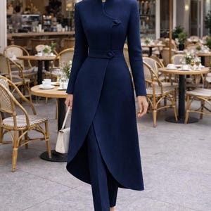 May include: A navy blue, long-sleeved coat dress with a wrap-style skirt and matching pants. The coat features a high collar and a single button closure at the waist. A small, white handbag and nude heels complete the outfit.