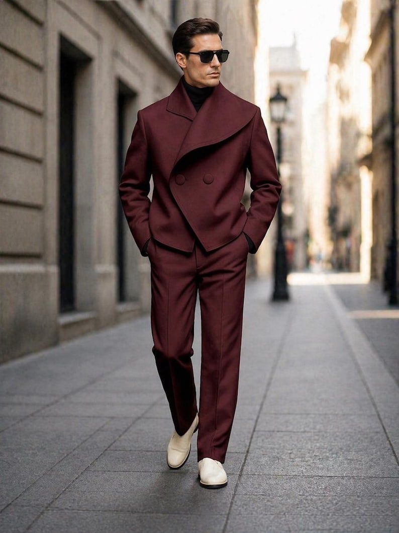 May include: A burgundy suit with a unique asymmetrical lapel design. The double-breasted jacket and matching trousers are paired with a black turtleneck and white loafers. The suit is a stylish ensemble for formal occasions.
