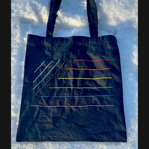 Op de afbeelding: Zwarte tote bag met een regenboogkleurig ontwerp geborduurd op de voorkant. Het ontwerp is een reeks parallelle lijnen die een driehoeksvorm vormen.