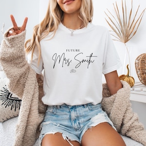 May include: White t-shirt with the text "FUTURE Mrs. Smith" in black script. The shirt also features a small ring graphic. The model is wearing denim shorts and a beige cardigan.