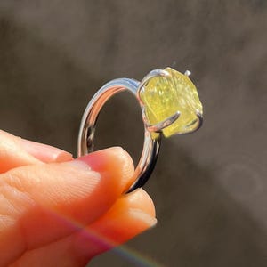 Op de afbeelding: Een zilveren ring met een grote, ruw geslepen, gele edelsteen. De edelsteen wordt vastgehouden door vier klauwen. De ring staat tegen een donkere achtergrond, met een regenboog in de rechterbenedenhoek.