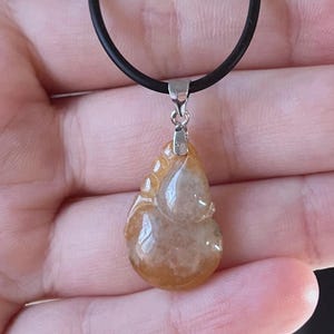 May include: A pendant necklace featuring a teardrop-shaped, amber-colored stone with a silver-tone bail. The stone is suspended from a black cord necklace. The pendant has a smooth, polished surface.