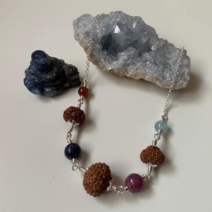 Può includere: Una collana a catena d'argento con varie perline, tra cui pietre preziose marroni, rosse, blu e viola. Sono presenti anche una statuina scolpita blu scuro e una grande formazione di cristalli azzurri.