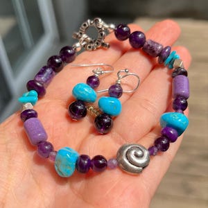 May include: A handmade jewelry set featuring a bracelet and matching earrings. The bracelet and earrings are adorned with purple amethyst beads, turquoise stones, and silver accents. The bracelet has a silver spiral charm and a toggle clasp.