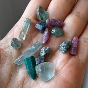May include: A collection of small, raw gemstones in various shapes and sizes. The assortment includes translucent light blue crystals, opaque dark green pointed stones, deep pink elongated pieces, and small, textured dark blue and green rocks.