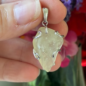 May include: A pale yellow pendant with a silver bail and decorative silver wire accents. The pendant is an irregular shape, possibly a natural stone or crystal. The background features colourful flowers.