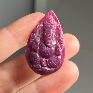 May include: A carved, teardrop-shaped pendant in a deep ruby color. The pendant features a detailed carving of a deity with multiple arms and an elephant head. The carving is intricate, with visible details of the deity's form.