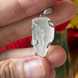 Könnte beinhalten: Ein klarer Quarzanhänger mit einer silberfarbenen Metalleinfassung. Der Quarz hat eine raue, unregelmäßige Form, und das Metall umschließt ihn in einem dekorativen Design. Der Anhänger wird in einer Hand gehalten.