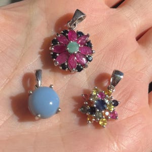 Op de afbeelding: Drie edelsteenhangers. Eén hanger heeft de vorm van een bloem met roze, zwarte en groene stenen. Een andere is een lichtblauwe ronde steen. De derde hanger heeft de vorm van een bloem met verschillende gekleurde stenen.