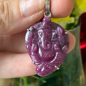 Peut inclure: Pendentif magenta foncé sculpté représentant la divinité hindoue Ganesha. Le pendentif est suspendu à une bélière argentée et présente une sculpture détaillée du dieu à tête d'éléphant. Le pendentif est tenu dans une main.