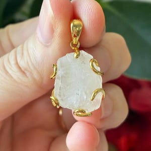 May include: A raw, clear, translucent crystal pendant encased in a polished gold-tone metal setting. The irregular, jagged stone is secured by four curved metal prongs and features a gold-tone bail at the top for attachment to a necklace chain.