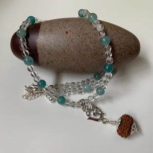 Può includere: Una collana con perline trasparenti e turchesi, drappeggiata su una pietra liscia di forma ovale. La collana ha una chiusura argentata e un ciondolo con una perla marrone strutturata. Le perline hanno un diametro di circa 0,8 cm.