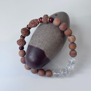 Può includere: Un braccialetto di perline con una miscela di materiali naturali. Il braccialetto include perline di legno marrone, perline di cristallo trasparente e perline marroni strutturate con accenti color oro. Il braccialetto è esposto su una pietra grigia e marrone.