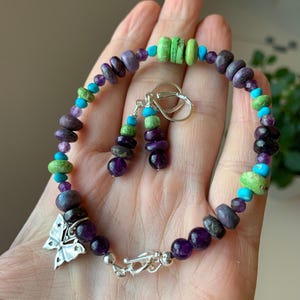 May include: A beaded bracelet and matching drop earrings featuring purple amethyst, bright green stone, and turquoise blue beads. The bracelet includes a silver-toned metal butterfly charm and a toggle clasp, while the earrings feature silver-toned hooks.