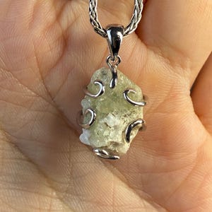 May include: A silver-toned necklace featuring a raw, light green gemstone pendant. The stone is encased in a decorative silver wire design and hangs from a braided chain. The pendant has a natural, textured appearance, showcasing its unique, organic form.