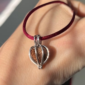Peut inclure: Un collier avec un pendentif argenté et un cordon bordeaux. Le pendentif est une cage contenant une pierre brune et texturée. Le collier est présenté sur un fond de couleur chair.