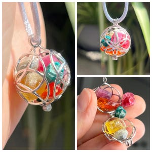 Puede incluir: Un collar con colgante plateado con un diseño de jaula esférica llena de piedras preciosas de colores. Las piedras preciosas incluyen tonos de rosa, naranja, amarillo y verde. El collar cuelga de un cordón blanco.