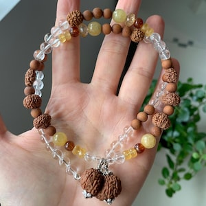 Peut inclure: Un collier à plusieurs rangs avec des perles en bois, des perles de cristal transparentes et des pierres précieuses jaunes et marron. Le collier a un pendentif avec deux pièces marron texturées. Le collier est tenu dans une main.