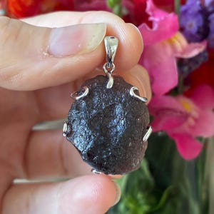 May include: A dark brown, irregularly shaped pendant with a silver bail and decorative silver accents. The pendant is held up against a blurred background of colourful flowers.