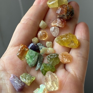 Peut inclure: Une collection de petites pierres précieuses brutes et multicolores de formes et de textures variées, incluant des nuances de jaune, vert, orange, violet et transparent, éparpillées dans une paume ouverte. Les pierres présentent des facettes naturelles.