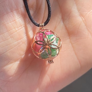 May include: A rose gold pendant necklace with a spherical cage design. The cage contains pink and green gemstones. The necklace has a black cord.