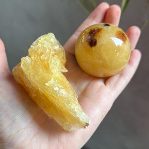 May include: A hand holds two golden-yellow crystals. One is a rough, textured cluster, and the other is a smooth sphere with brown markings. The crystals are illuminated, showcasing their natural color and form. The background is blurred.