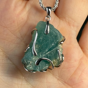 Op de afbeelding: Een turquoisekleurige, ruwe edelsteen hanger met een zilverkleurige draadwikkeling en balein. De hanger hangt aan een zilverkleurige ketting. De edelsteen heeft een ruwe, natuurlijke textuur.