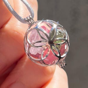 May include: A silver pendant necklace with a spherical cage design. The cage contains a pink and green stone. The necklace hangs from a light grey cord.