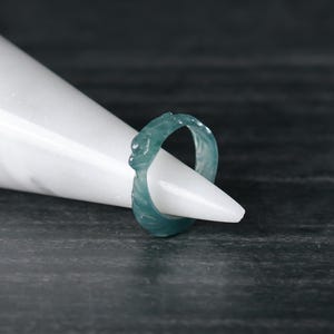 May include: A teal-colored ring with a carved design, displayed on a white cone against a dark background. The ring's intricate details are visible, showcasing its unique texture and shape. The ring appears to be made of jade or a similar stone.