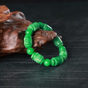 May include: A jade bracelet with a mix of round and cylindrical beads. The bracelet is a vibrant green color and is displayed on a piece of dark wood.