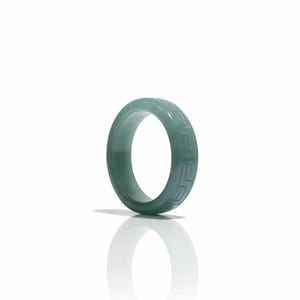 May include: A jade green ring with a geometric pattern carved around the band. The ring is a solid color and reflects light, with a shadow cast on the white surface.