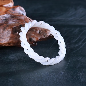 May include: A white, twisted bracelet with a braided design. The bracelet is made of a smooth, opaque material and is displayed on a dark surface with a wooden accent.