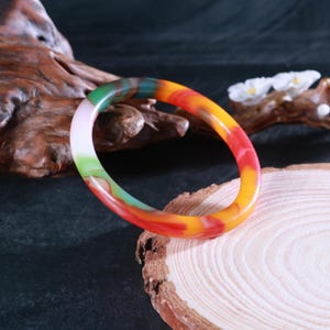 May include: A colorful bangle bracelet with a marbled design. The bracelet features a blend of colors, including green, red, orange, and yellow. The bracelet is displayed on a wooden surface.