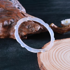 May include: A translucent, light blue jade bangle bracelet. The bracelet has a smooth, polished finish and a delicate, carved design. The bracelet is displayed on a wooden surface with a natural, organic texture.