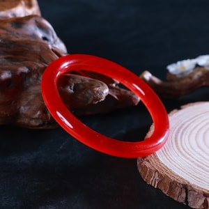 May include: A bright red bangle. The circular bracelet is made of a translucent material and is displayed on a piece of wood against a dark background. The bangle's smooth surface reflects light, showcasing its rich colour and simple design.