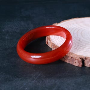 May include: A vibrant red bangle bracelet. The circular bracelet is made of a translucent material, and is displayed on a dark surface with a wooden slice in the background. The bangle is a piece of jewellery, and is a circular shape.