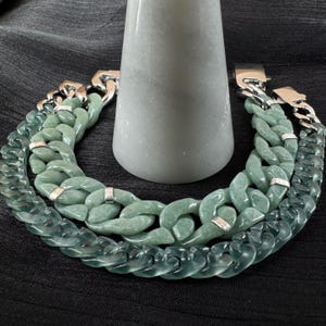 May include: Two jade-colored chain link bracelets with silver clasps are displayed. The bracelets are made of large, interlocking links. The bracelets are arranged in a semi-circle around a light gray, cylindrical object.