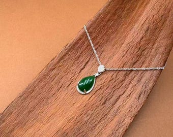 May include: A silver necklace with a teardrop-shaped green jade pendant. The pendant is surrounded by a silver frame and has a small heart-shaped accent. The necklace is displayed on a wooden surface.