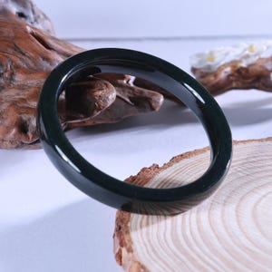 May include: A dark green jade bangle bracelet. The smooth, polished bracelet is circular and appears to be made of a solid, opaque material. The bracelet is displayed on a wooden surface.