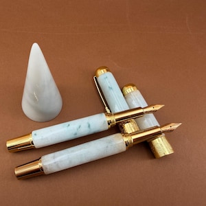 May include: Two fountain pens with gold accents and light green and white marbled barrels are displayed. A white marble cone-shaped pen holder is to the left. The pens have gold nibs and caps. The background is a solid brown color.