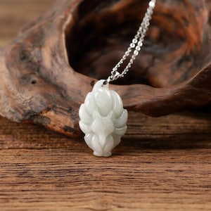 May include: A light green jade pendant in the shape of a fox, suspended from a silver chain. The fox pendant is intricately carved with detailed features. The background is a wooden surface.