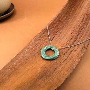 May include: A green jade donut pendant necklace with a silver chain. The pendant is a circular shape with a twisted design. The necklace is displayed on a wooden surface with a warm, brown tone.