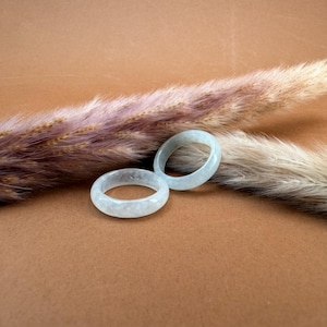 May include: Two light green jade rings are displayed on a brown surface, with dried ornamental grass in the background. The rings have a smooth, polished finish and a circular design. The rings are a light, translucent green color.