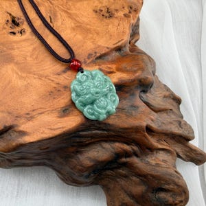 May include: A jade pendant necklace featuring a carved, light green pendant with intricate details. The pendant is suspended from a dark cord with a red bead accent. The necklace rests on a textured, brown wooden surface.