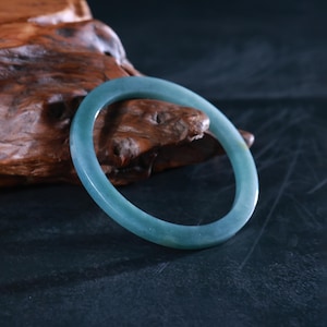 May include: A translucent, light teal jade bangle bracelet. The smooth, circular bracelet is displayed against a dark background and a piece of textured, brown wood. The jade has subtle color variations.