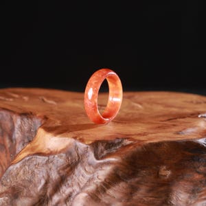 May include: A vibrant orange-red ring, likely jade or a similar gemstone, stands out against a dark background. The ring's smooth surface reflects light, showcasing its rich color and natural variations. The ring is displayed on a wooden surface.