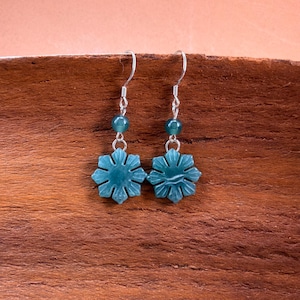 May include: A pair of teal snowflake earrings with silver hooks. Each earring features a carved snowflake pendant and a teal bead. The earrings are displayed on a wooden surface.