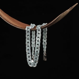May include: A light blue chain necklace with silver-colored accents hangs from a curved wooden display. The necklace features a chunky link design and a silver clasp. The background is solid black.