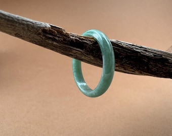 May include: A light green jade bangle bracelet. The smooth, circular bracelet is displayed on a piece of weathered wood against a neutral background. The jade has a translucent quality, showcasing its natural color and texture.